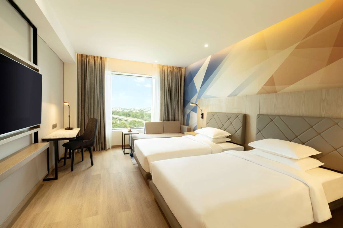 A hotel room with two beds, a desk with a chair, a TV, and a large window with curtains.