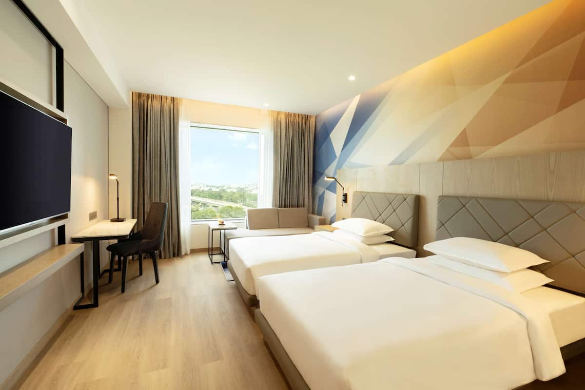 Habitación de hotel con dos camas, un escritorio con silla, un televisor y un ventanal con cortinas.