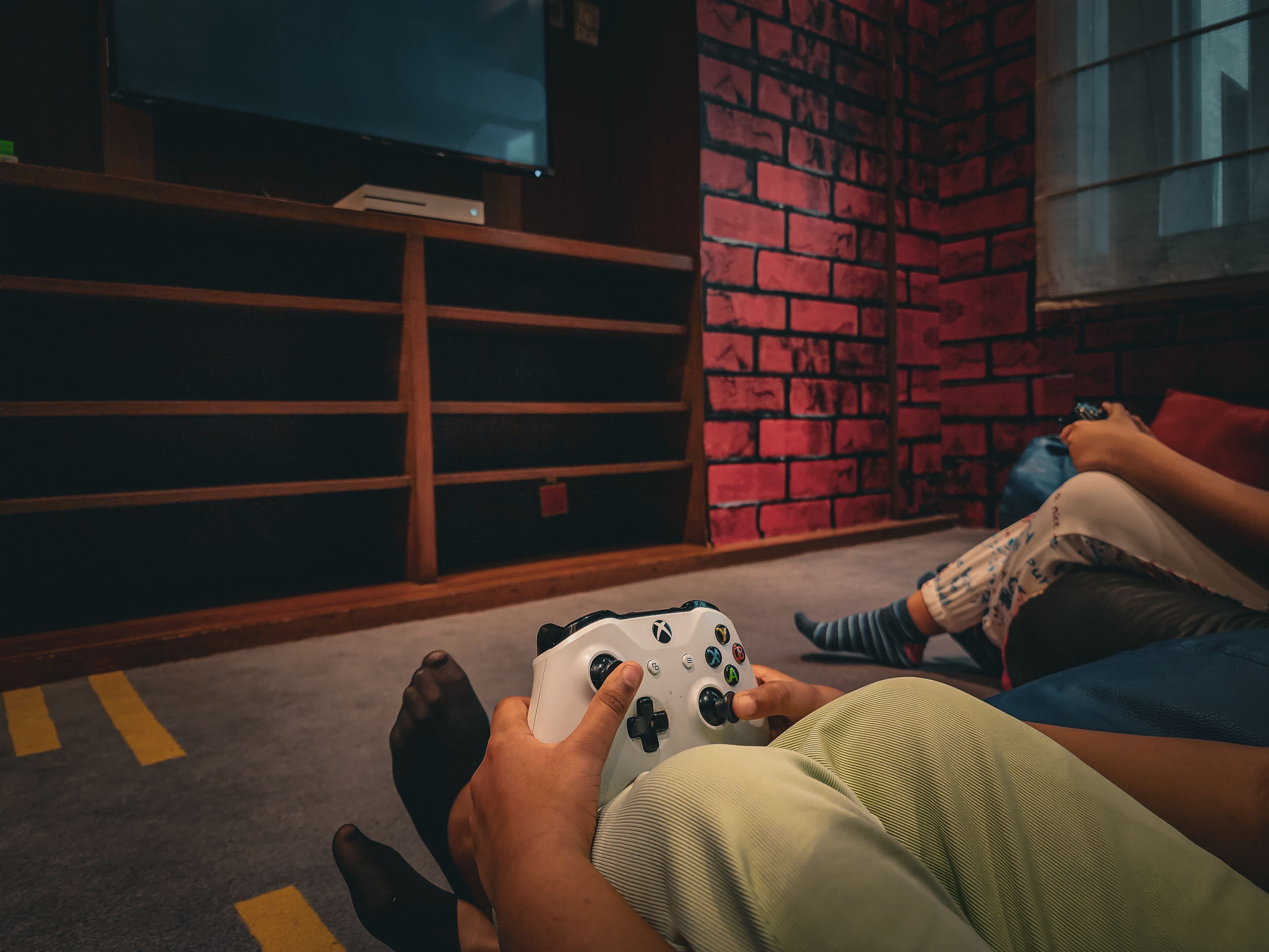 Une personne tenant une manette de jeu dans une pièce dont un mur est en briques et qui contient une étagère avec divers objets.