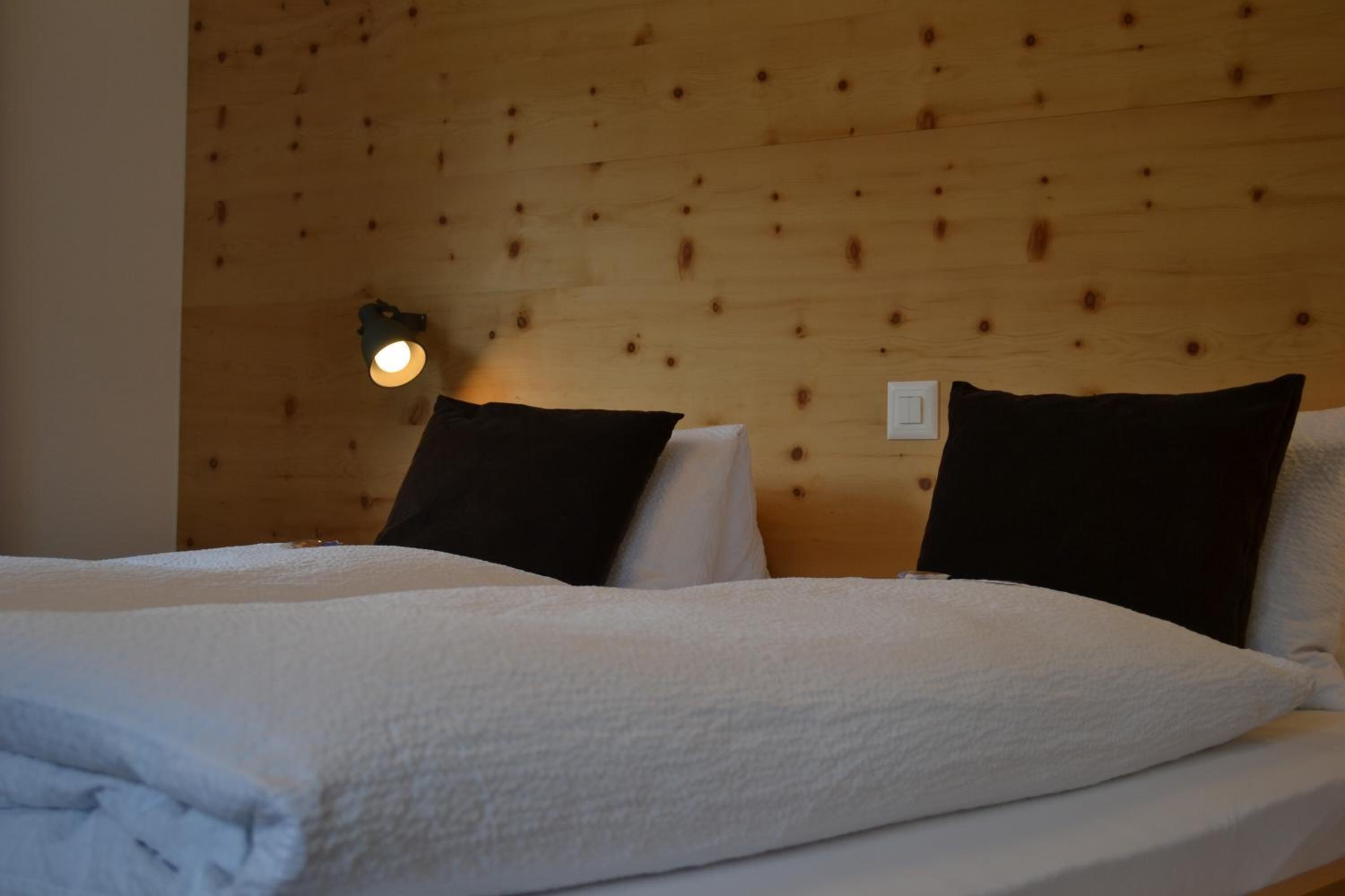 Un lit double avec du linge de lit blanc et deux oreillers foncés, une lampe fixée au mur et un mur recouvert de panneaux en bois.