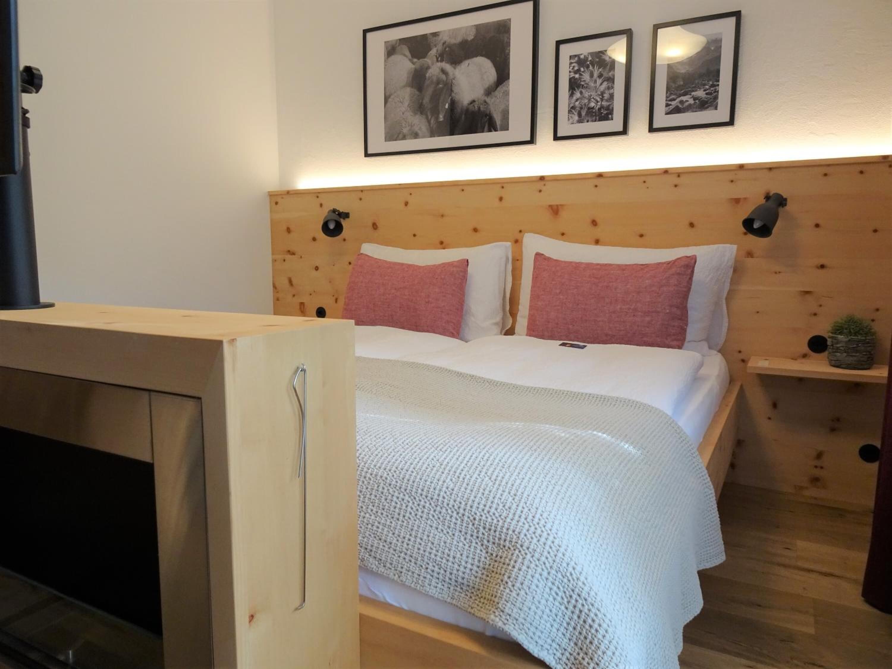 A bedroom with a wooden headboard, a bed with white bedding and pink pillows, a small bedside table with a lamp, and a wall-mounted shelf with a plant.