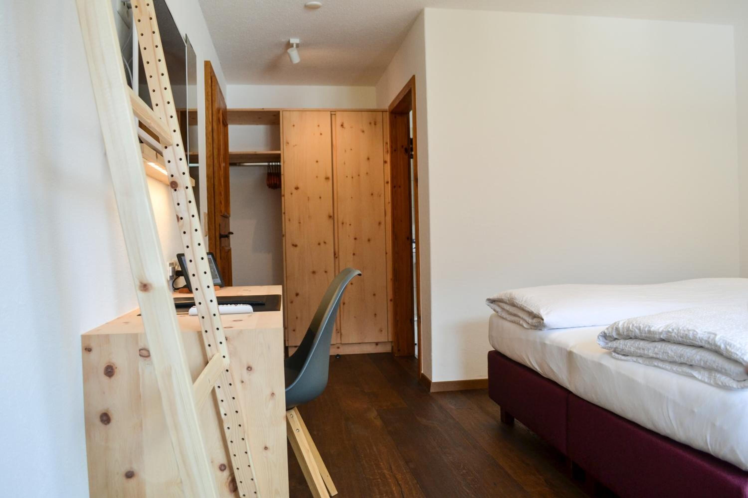 Une chambre à coucher avec un lit, un bureau et une chaise. On y trouve également une armoire et une porte qui donne sur une autre pièce.
