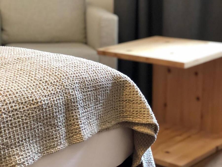 Una cama con una colcha texturizada, una mesita de noche de madera y un sofá en una habitación con una pared lisa y una cortina.