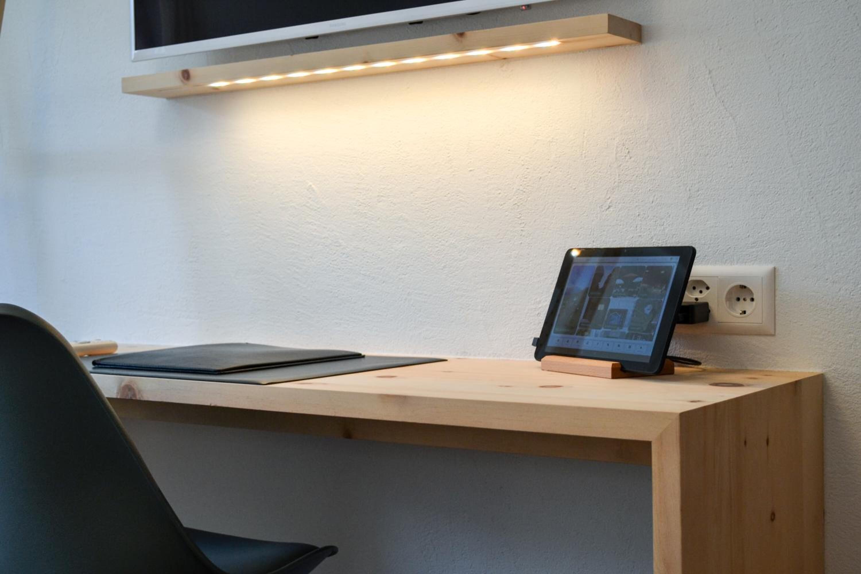 Ein Holztisch mit einem Tablet, ein schwarzer Stuhl und ein Wandregal über einer Steckdose.