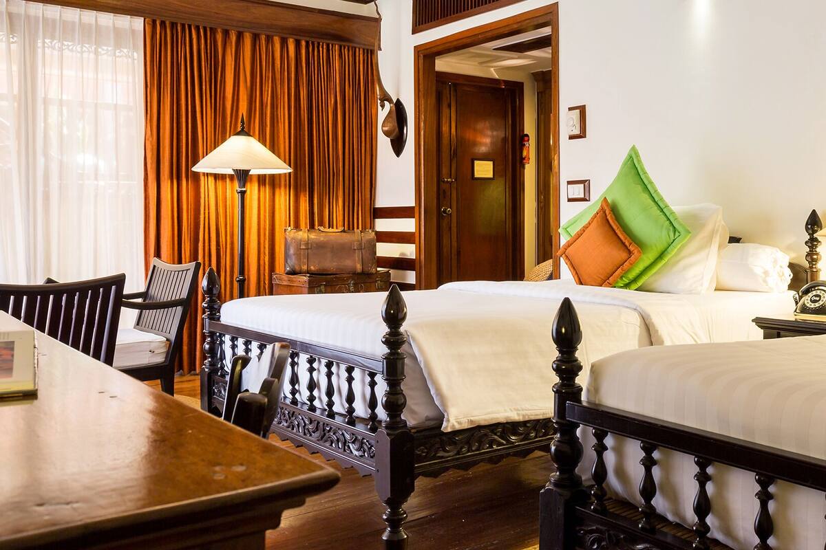 A hotel room with two beds, a wooden table, a chair, a lamp, and a suitcase.