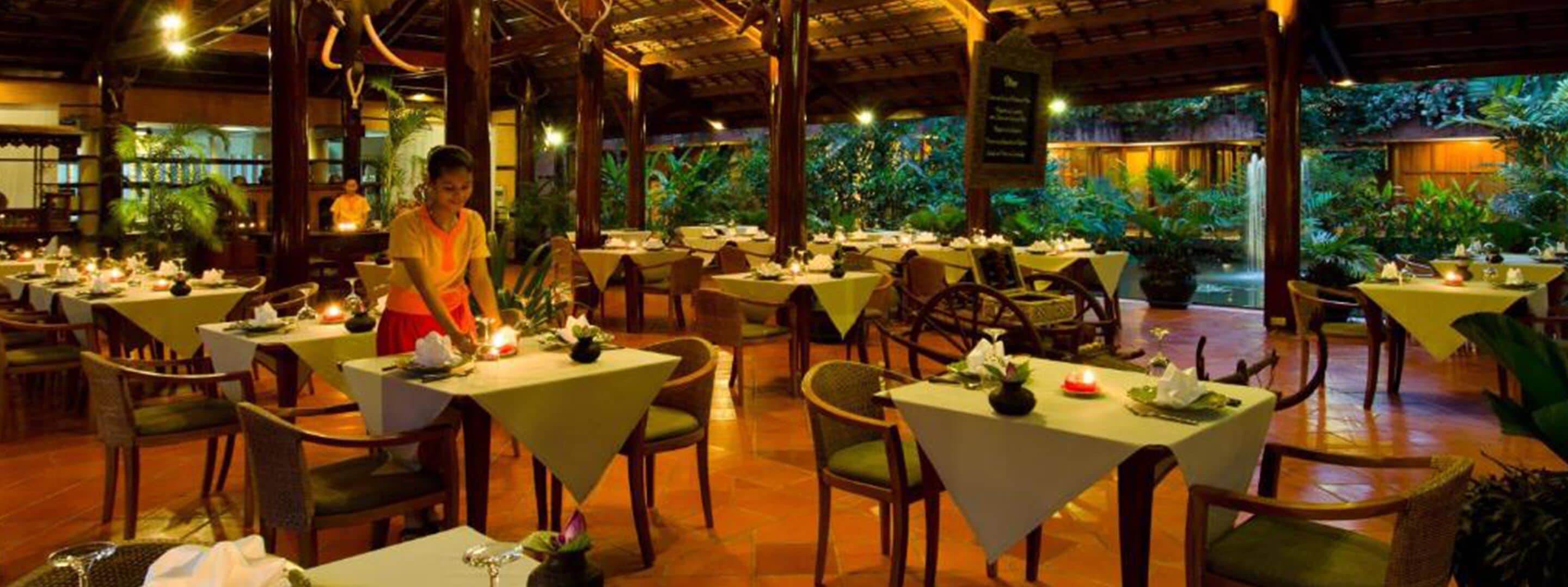 Un restaurante con varias mesas dispuestas para comer, un camarero atendiendo a los clientes y un exuberante jardín interior al fondo.
