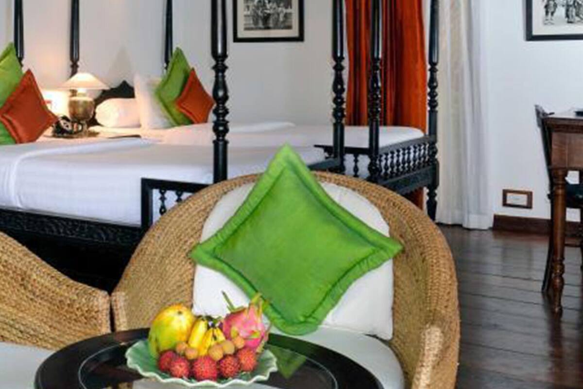 A hotel room with a bed, two wicker chairs with green cushions, a table with fruit, and a wooden floor.