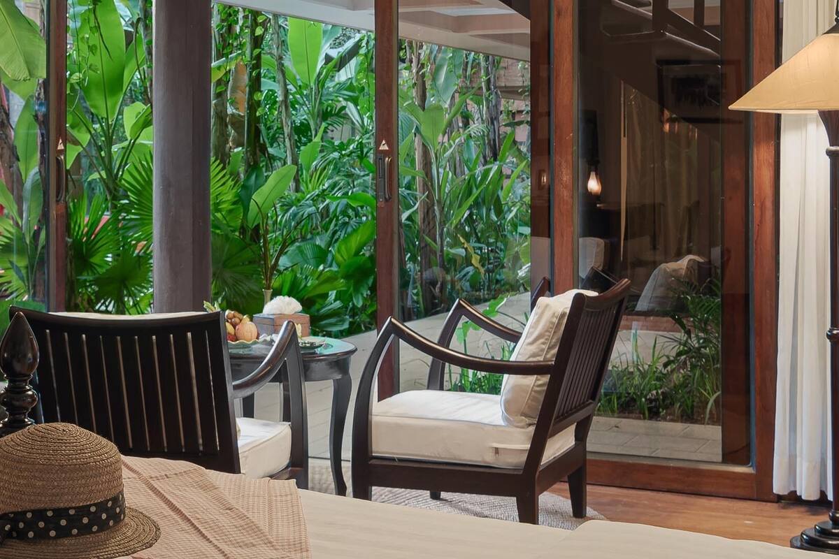 A room with a view of lush greenery, a dining table with chairs, a lamp, and a hat on the table.