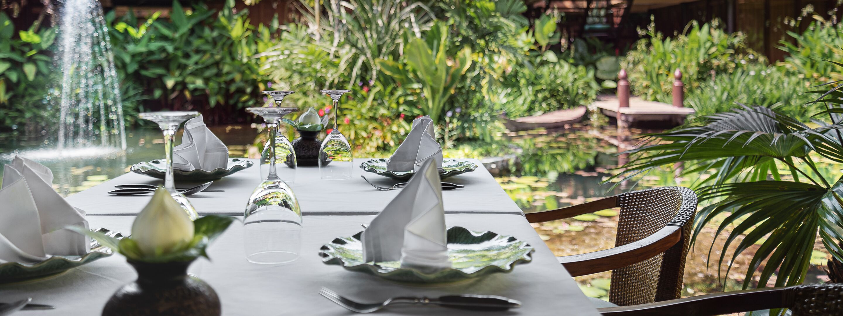 Una mesa dispuesta con cristalería transparente y platos, rodeada de exuberante follaje y una fuente de agua.