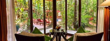 A room with a view of an outdoor garden area, featuring a table with a fruit bowl and two chairs.
