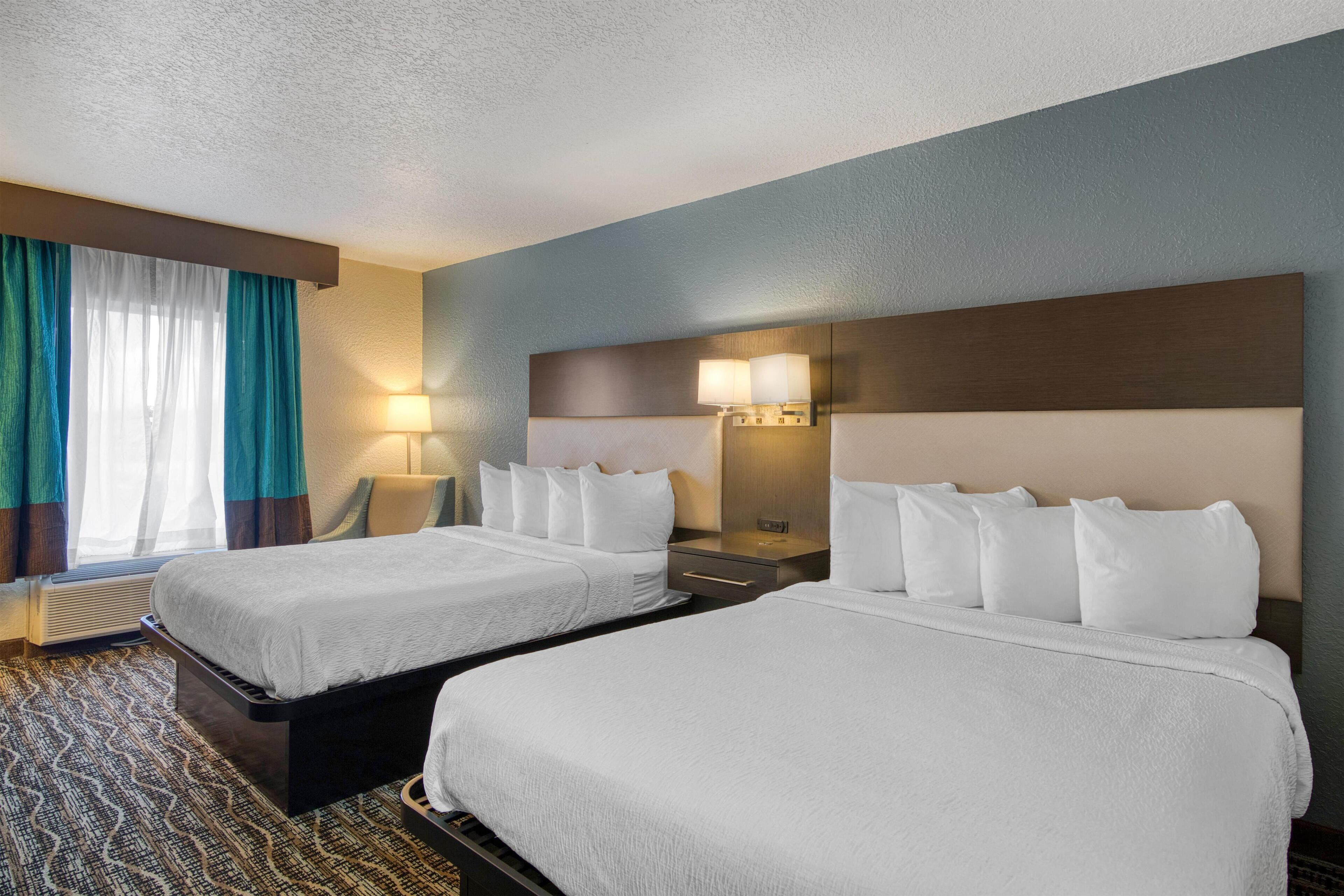 A hotel room with two beds, a nightstand with a lamp, a headboard with a light, and a window with curtains.