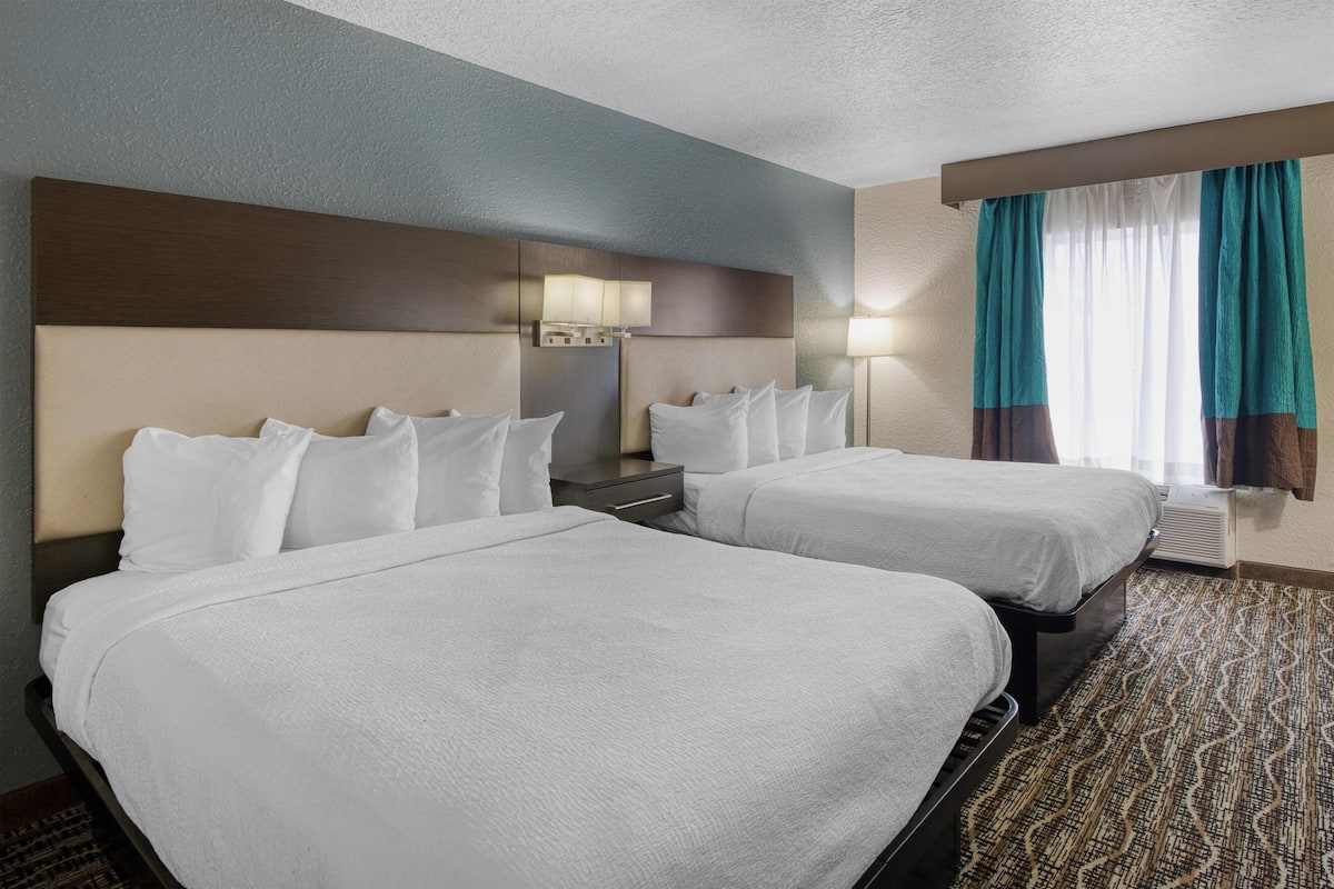 A hotel room with two beds, a nightstand, a lamp, and a window with curtains.