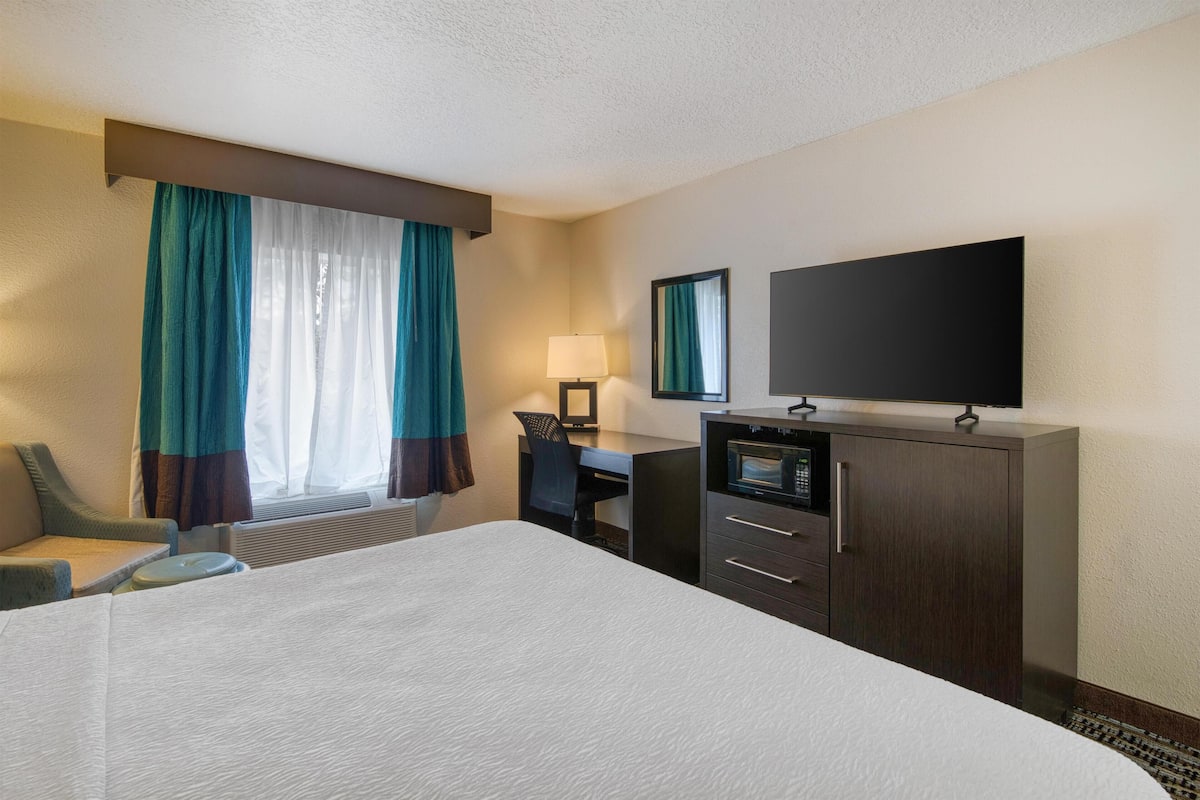 A hotel room with a large bed, a desk with a computer, a television, and a window with curtains.