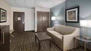 A hotel room with a patterned carpet, a sofa, a small table, a lamp, and a door with a notice. - Best Western Waldo Inn and Suites (Waldo)