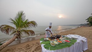 A beachside dining setup with a chef preparing food, a table covered with a white cloth, and a palm tree in the background.