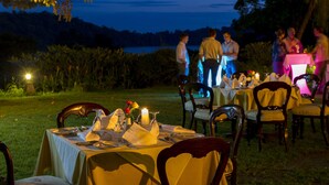 Uma área de refeições ao ar livre com mesas preparadas para uma refeição, rodeada por cadeiras e vista para um lago ao entardecer.