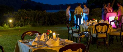 An outdoor dining area with tables set for a meal, surrounded by chairs and a view of a lake at dusk.