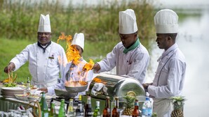 Cozinheiros preparando refeições ao ar livre, com uma churrasqueira e diversos utensílios de cozinha.