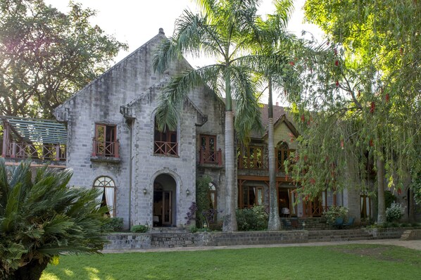 A historic stone building with a central arched doorway, a balcony with wooden railings, and a well-maintained garden with palm trees and shrubs. - Arusha Serena Hotel (Arusha)