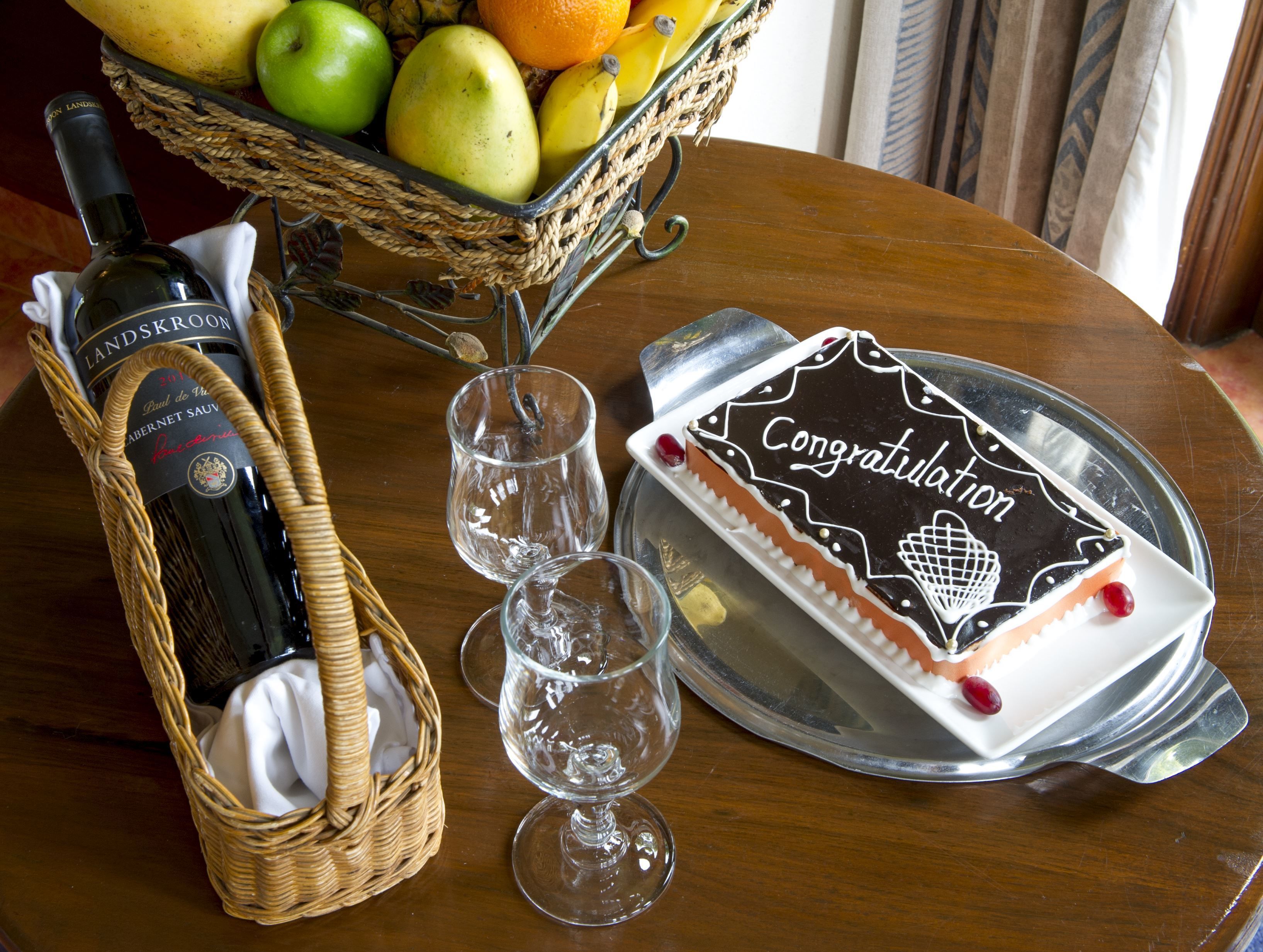 A table with a bottle of wine, a fruit basket, two wine glasses, and a congratulatory cake.