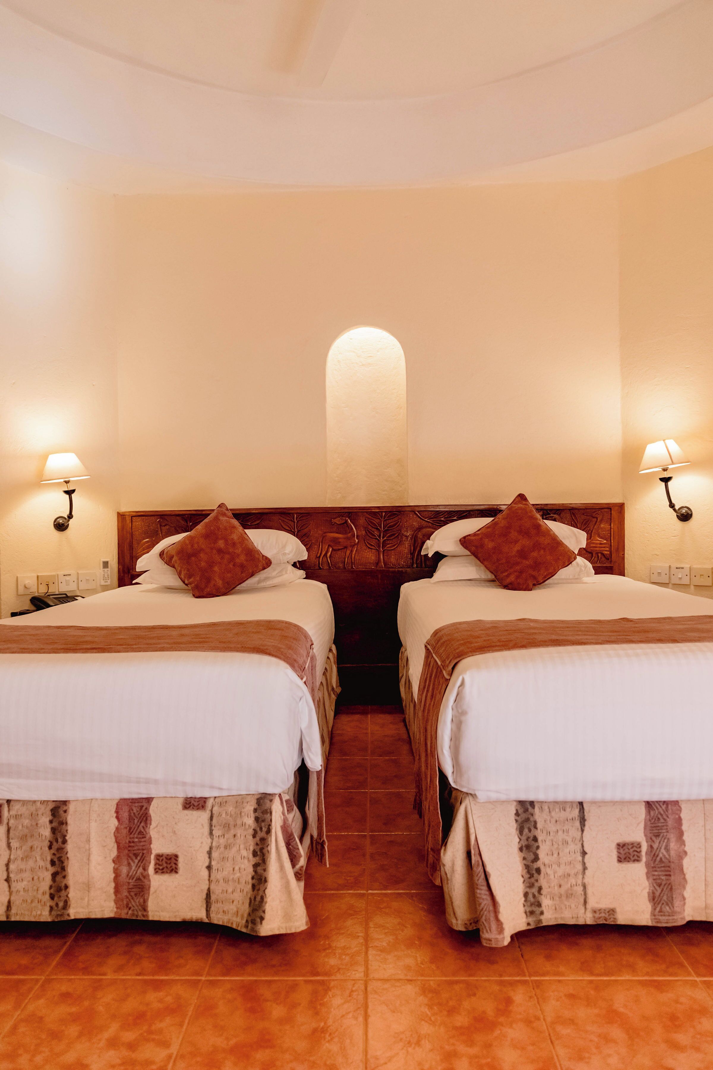 A hotel room with two beds, each with a wooden headboard, white bedding, and brown throw pillows. There are wall-mounted lamps on either side of the beds.