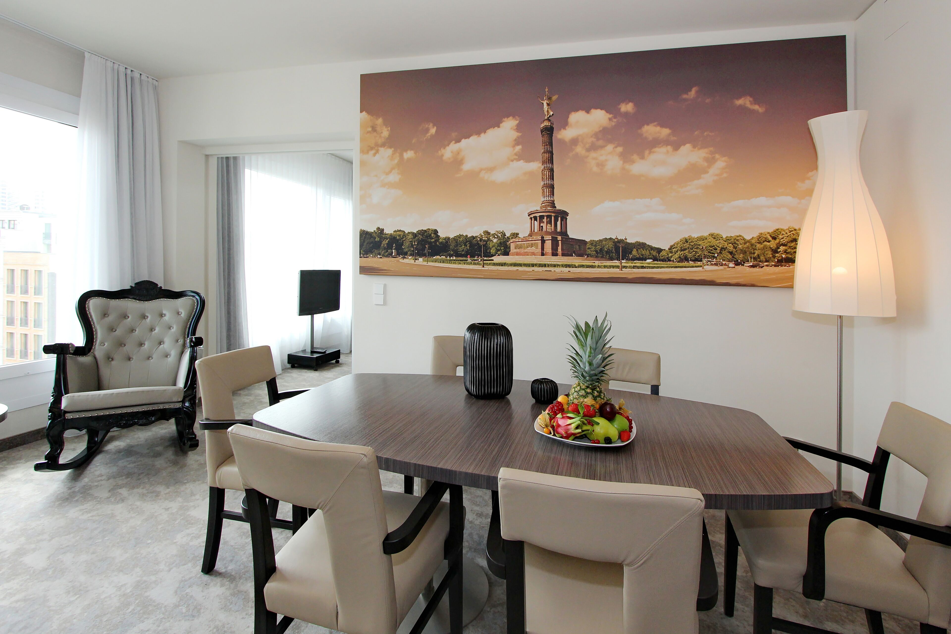 A hotel room with a round dining table, six chairs, a fruit bowl, a large painting of a tower, a lamp, and a classic armchair.