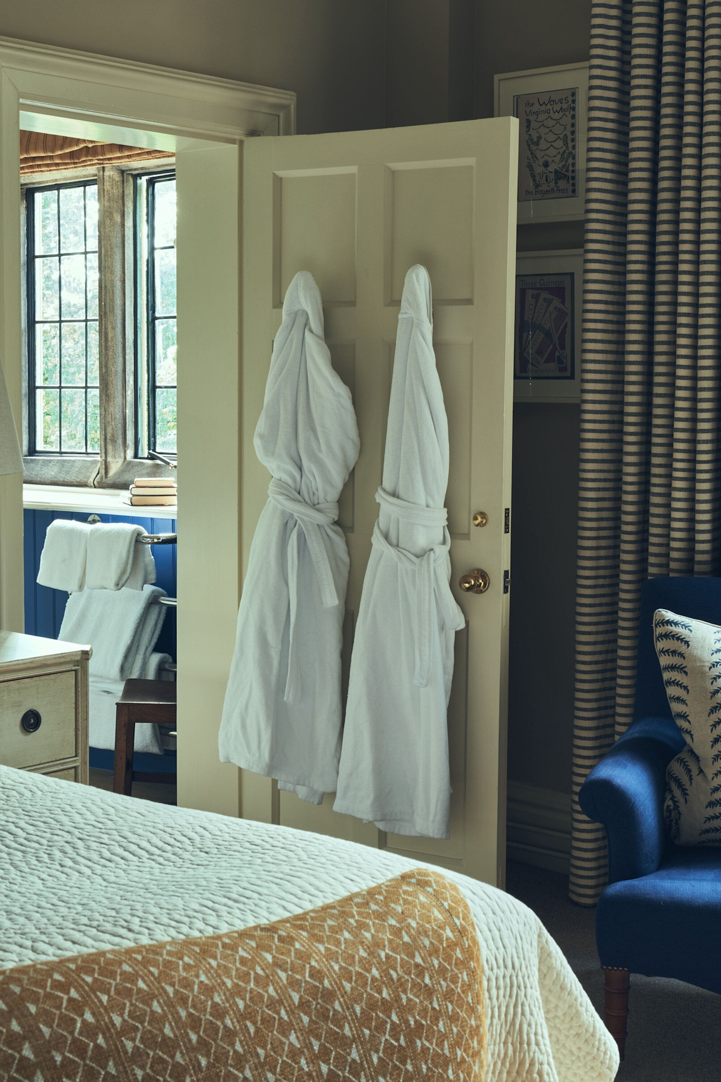 A room with a bed, a chair, and two white robes hanging on a door.