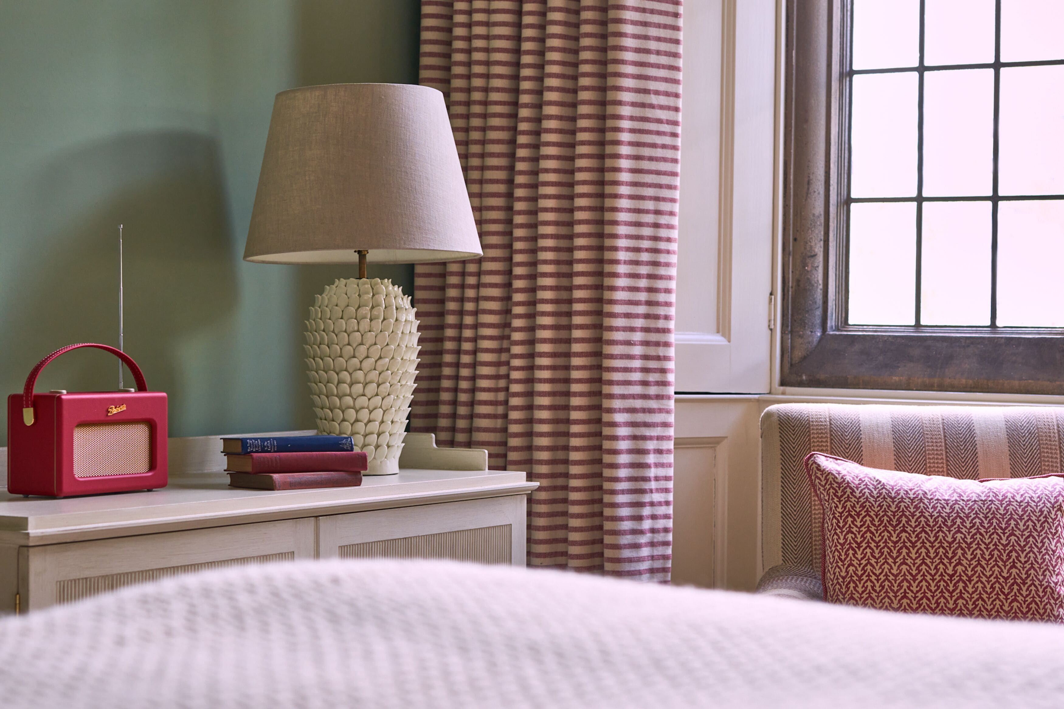 A neatly arranged bedroom with a bed, a bedside table with a lamp, a radio, books, and a decorative vase.