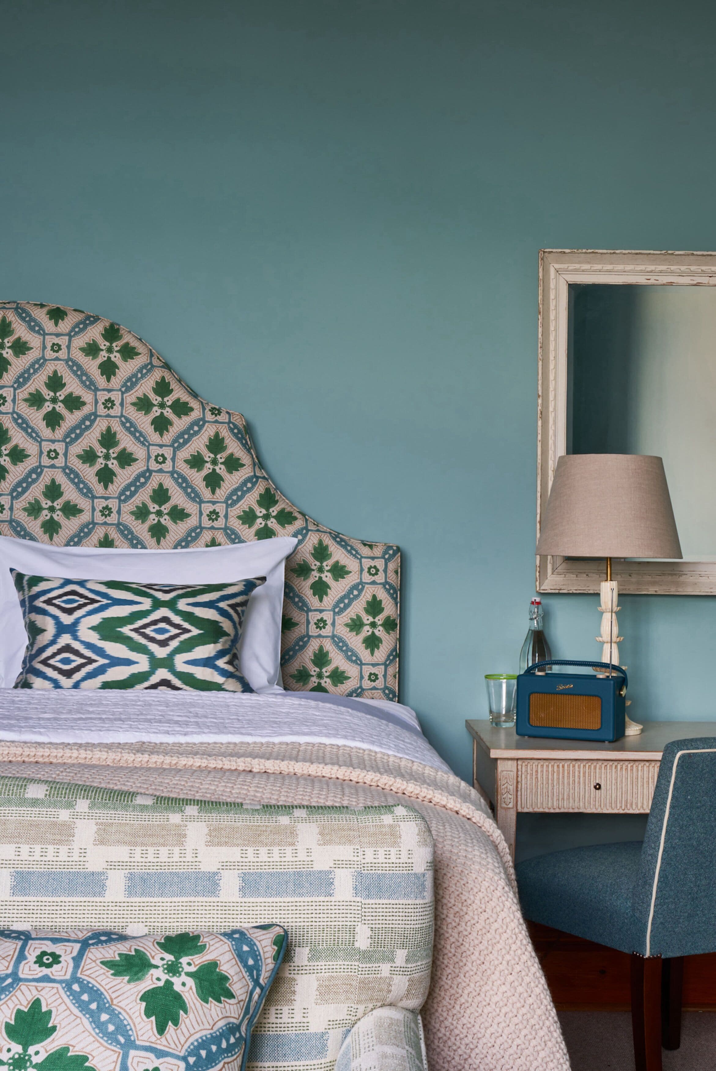 A bedroom with a patterned headboard, a bed with multiple pillows, a bedside table with a lamp, and a chair.