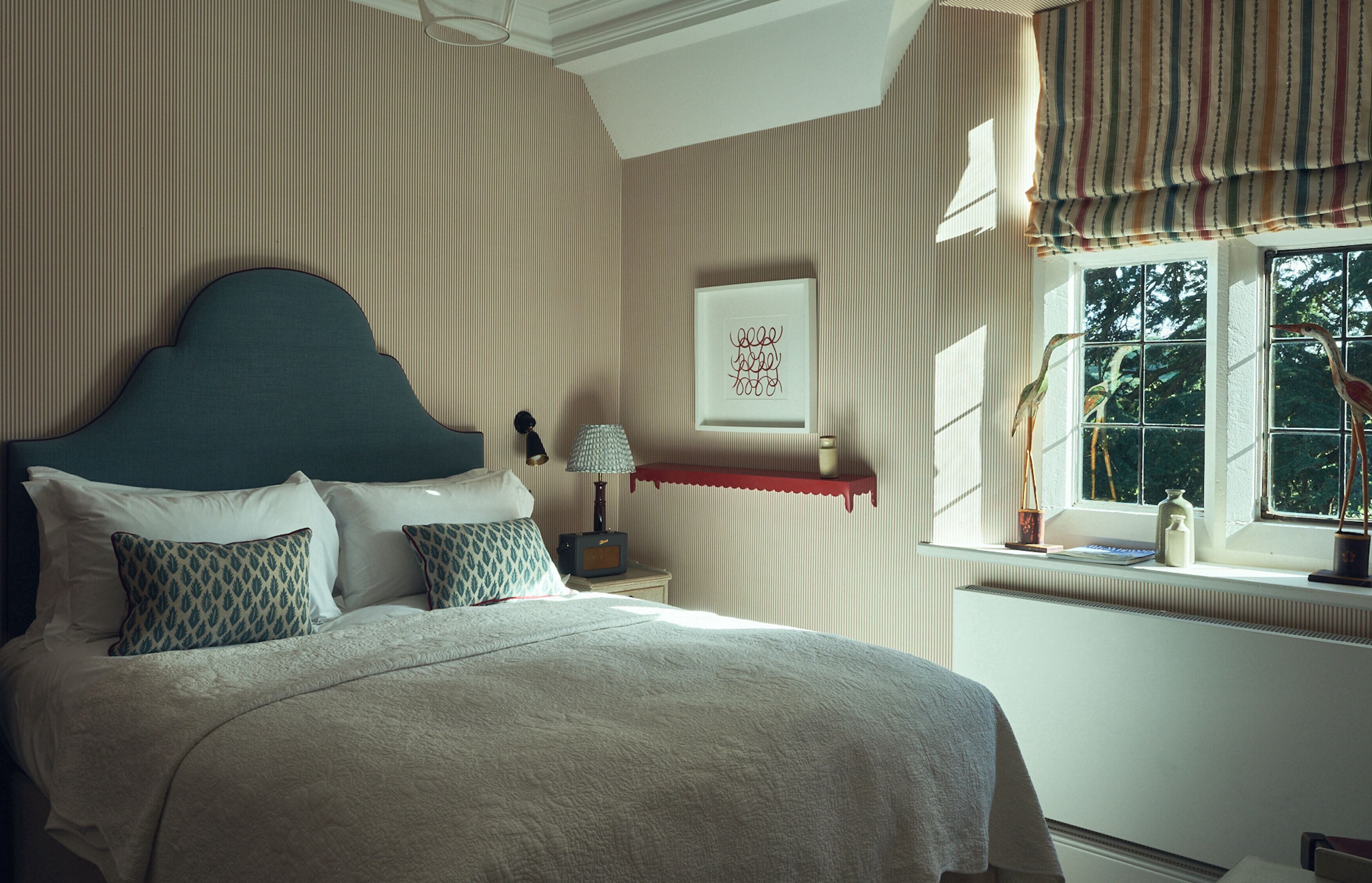 A bedroom with a large bed, a bedside table with a lamp, a framed picture on the wall, and a window with a view of trees.