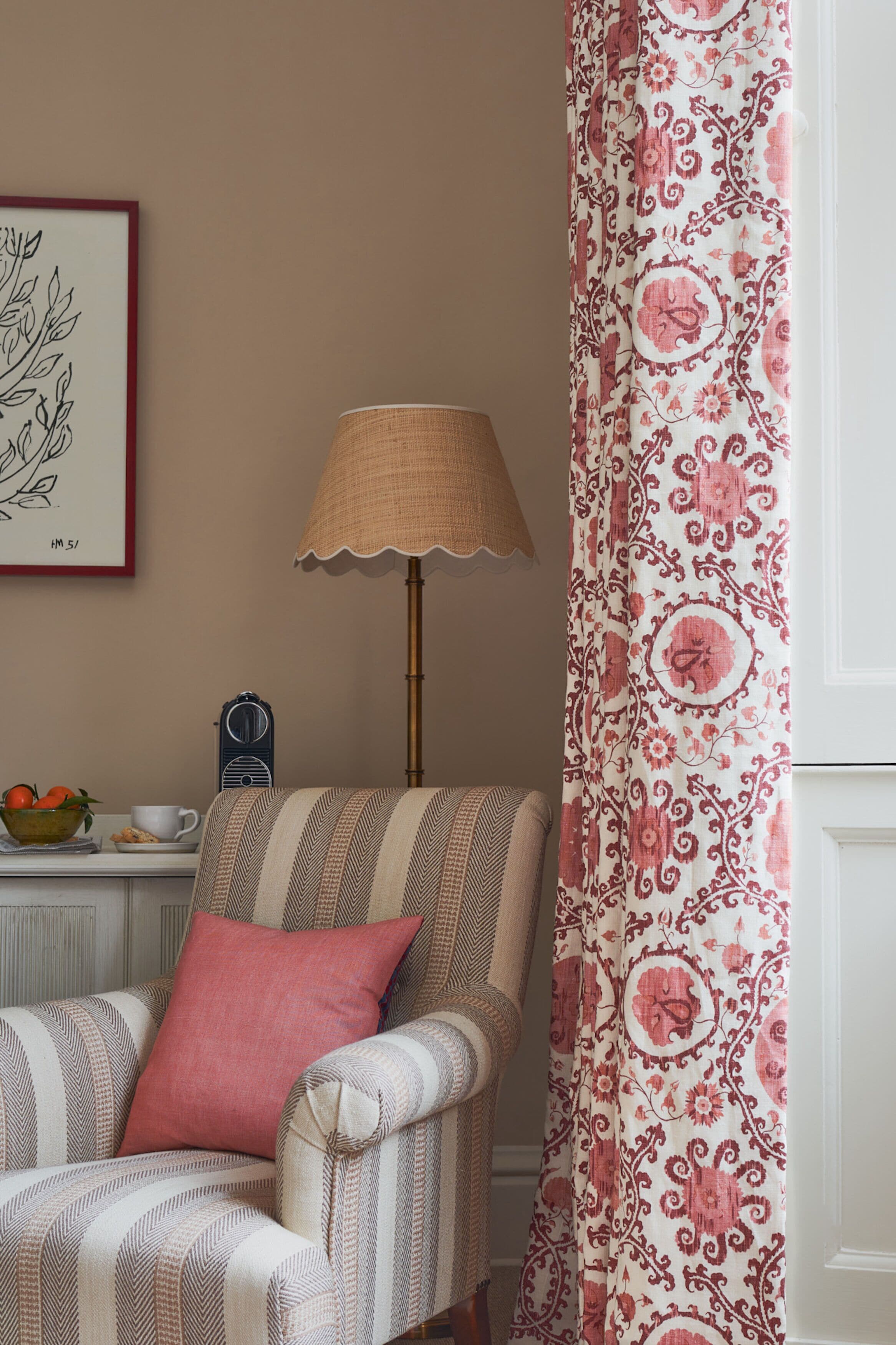 Una silla con estampado a rayas, un cojín rojo, una lámpara, una obra de arte enmarcada y una cortina estampada.