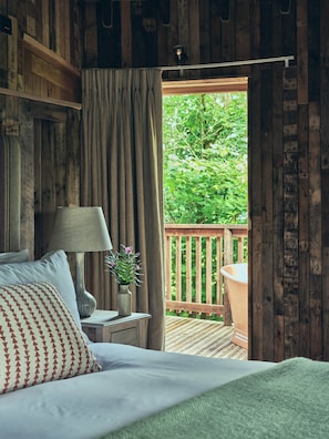A wooden room with a bed, a nightstand, a lamp, and a view of a wooden deck and greenery outside. - Wildhive Callow Hall (Ashbourne)