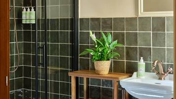 Un baño con mampara de vidrio, un estante de madera con una planta en maceta, un lavabo con grifo y una toalla colgada en un perchero.