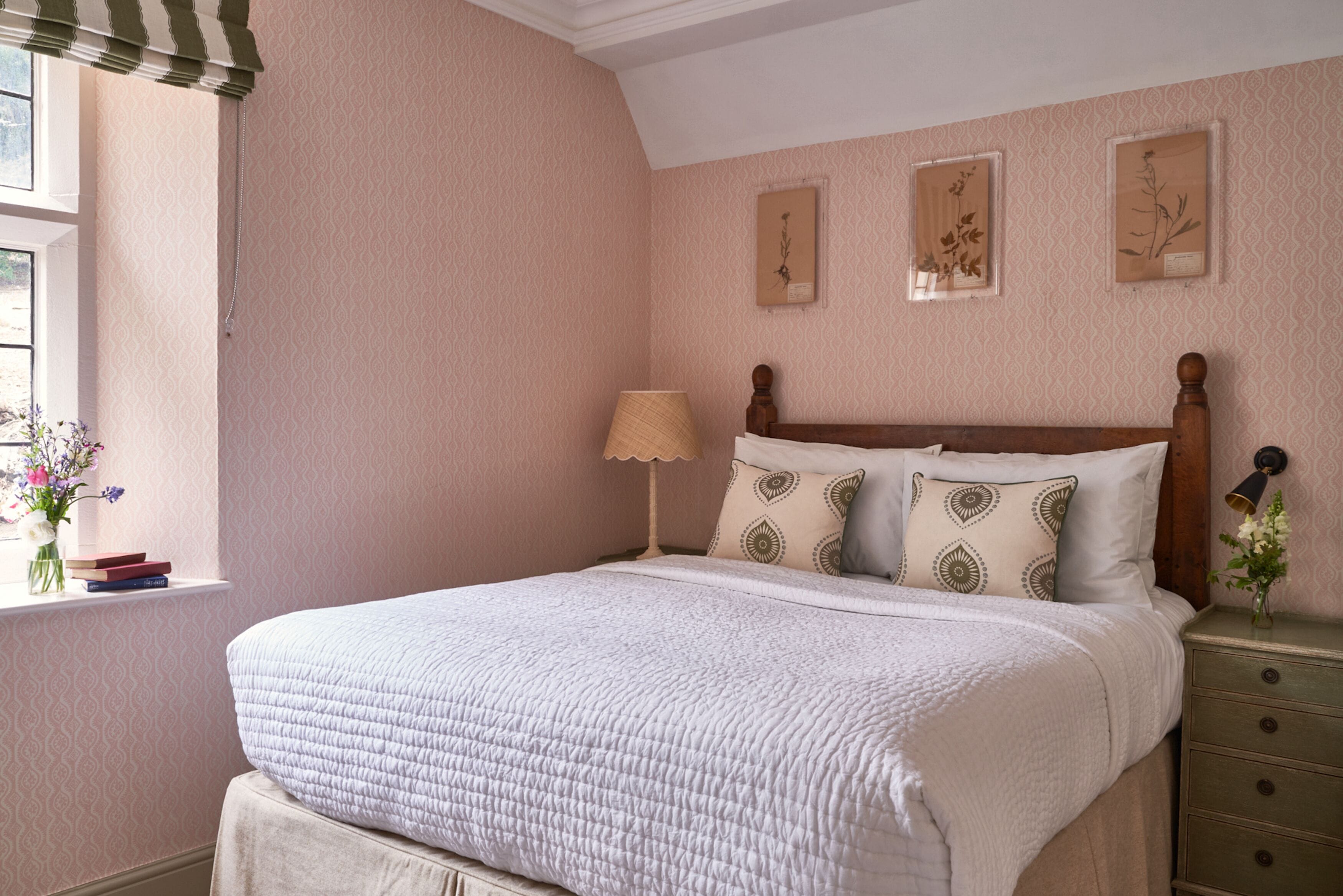 A neatly arranged bedroom with a large bed, a bedside table with a lamp, and framed botanical prints on the wall.