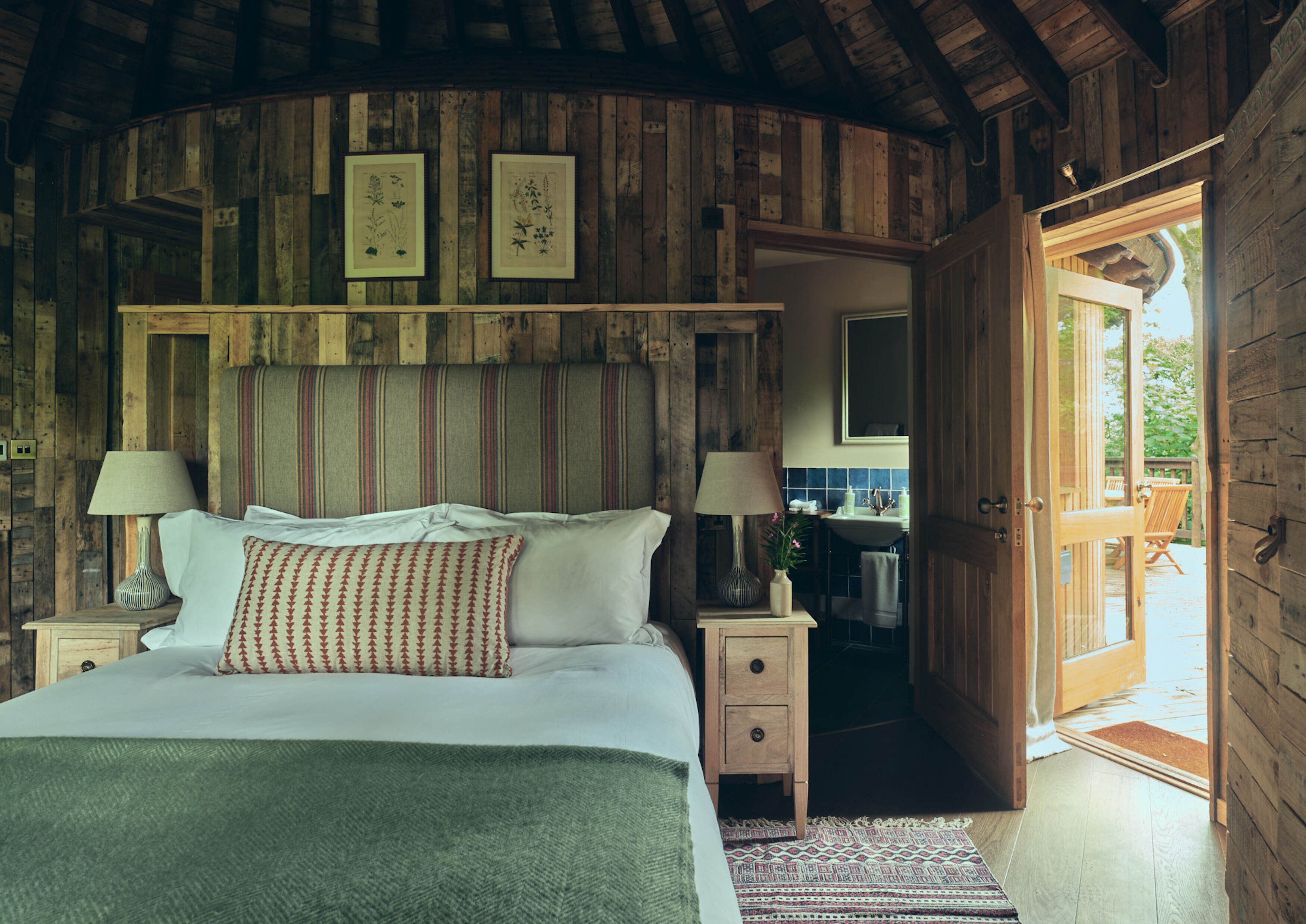 A rustic wooden bedroom with a bed, bedside lamps, a small bedside table, and a door leading to a bathroom and outdoor area.