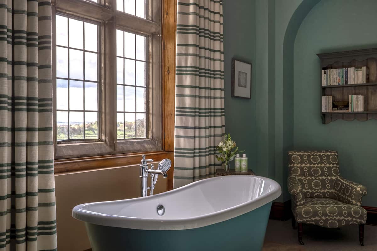 Ein Badezimmer mit einer freistehenden Badewanne, einem Holzstuhl, einem Bücherregal und einem Fenster mit Vorhängen.