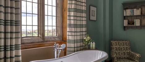 A bathroom with a freestanding bathtub, a wooden chair, a bookshelf, and a window with curtains.
