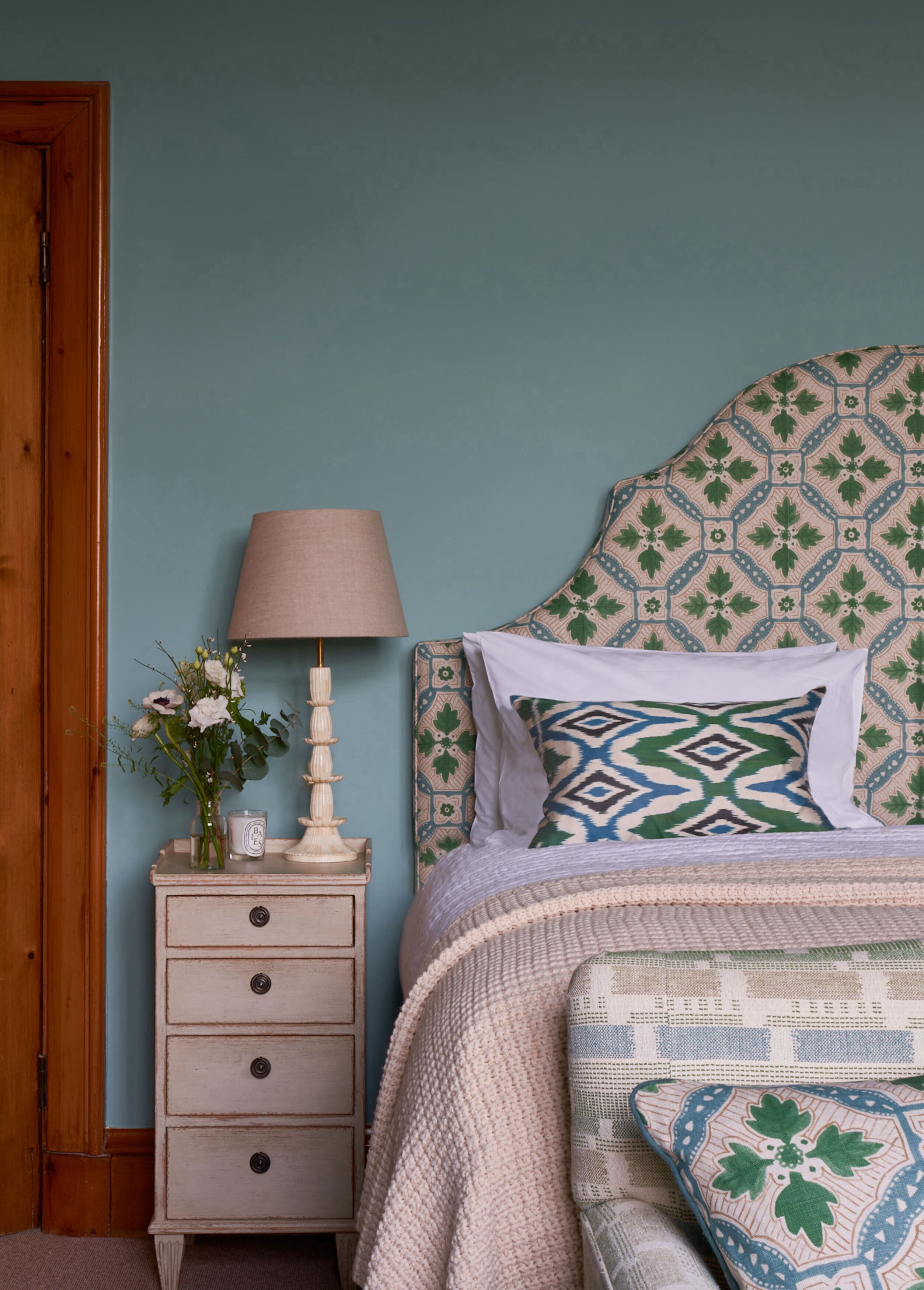 Un dormitorio con un cabecero estampado, una mesita de noche con lámpara y un arreglo floral.
