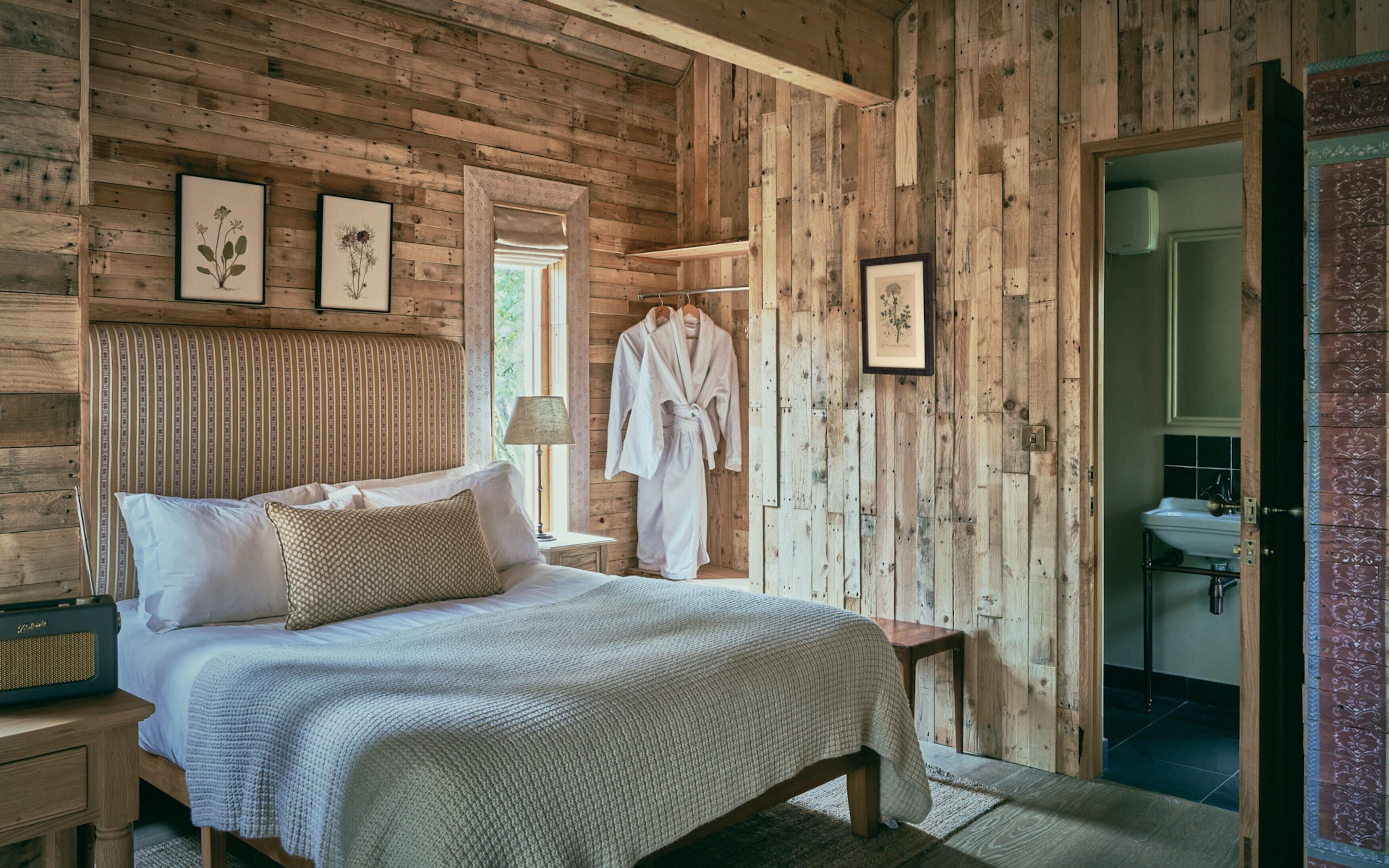 A wooden cabin bedroom with a bed, a nightstand, a robe, and framed botanical prints on the wall.