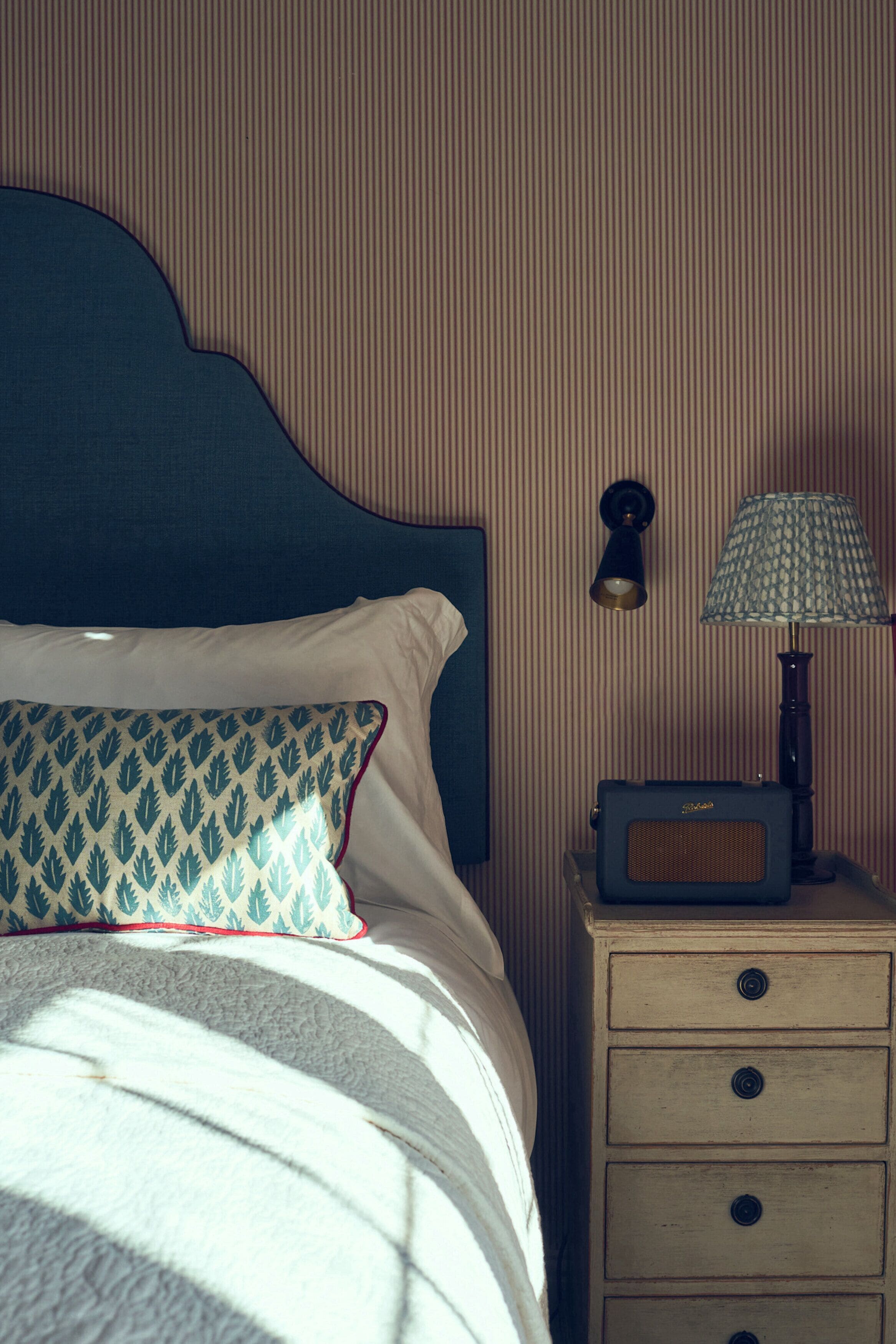 Una cama bien hecha con almohadones estampados, un cabecero, una lámpara de noche y una mesita de noche de madera con cajones.