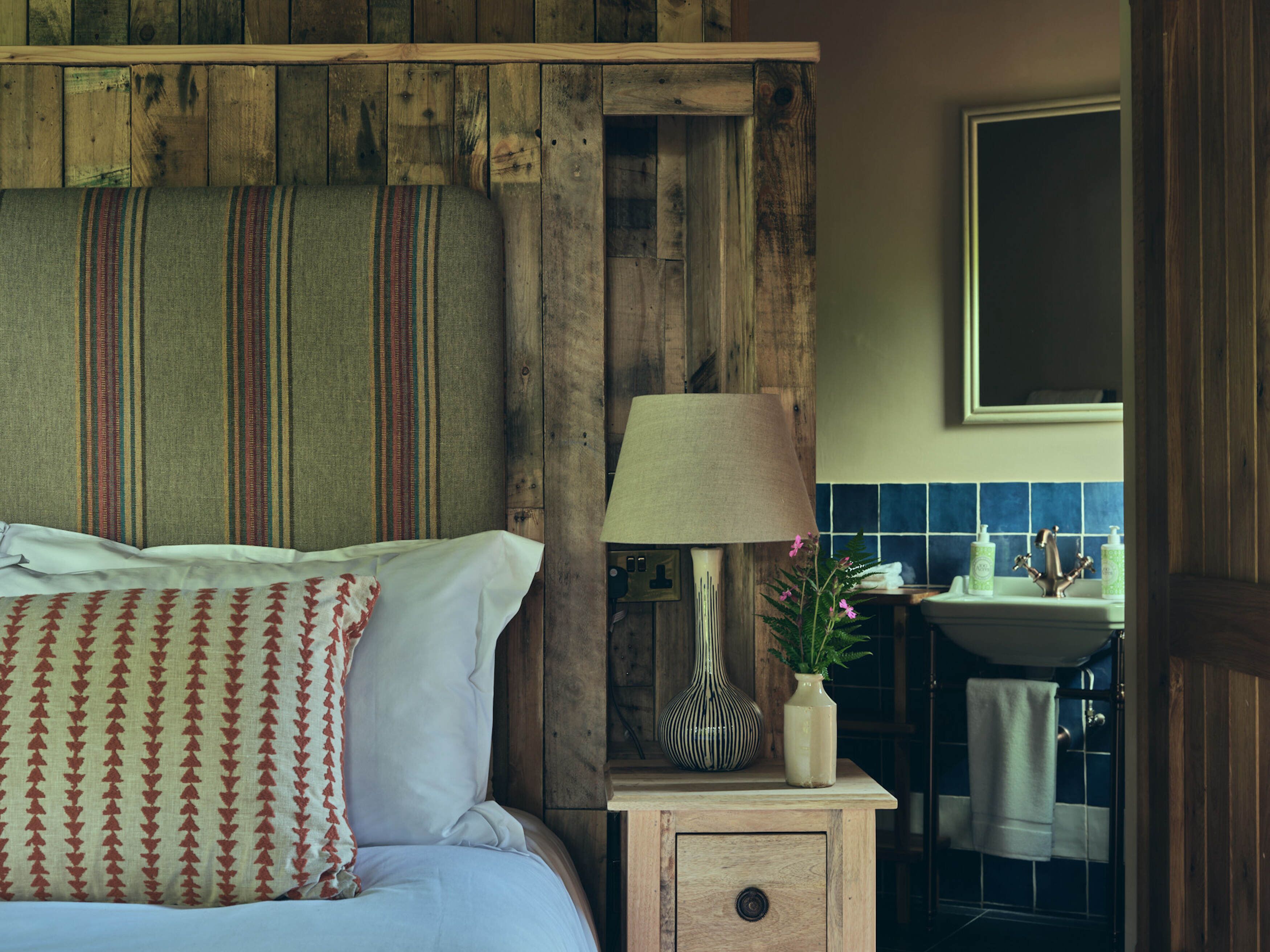 Un dormitorio rústico con cabecera de madera, una lámpara de noche, una mesita con un jarrón, y vistas a un baño con lavamanos y azulejos azules.