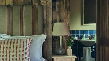 Un dormitorio rústico con cabecera de madera, una lámpara de noche, una mesita con un jarrón, y vistas a un baño con lavamanos y azulejos azules.
