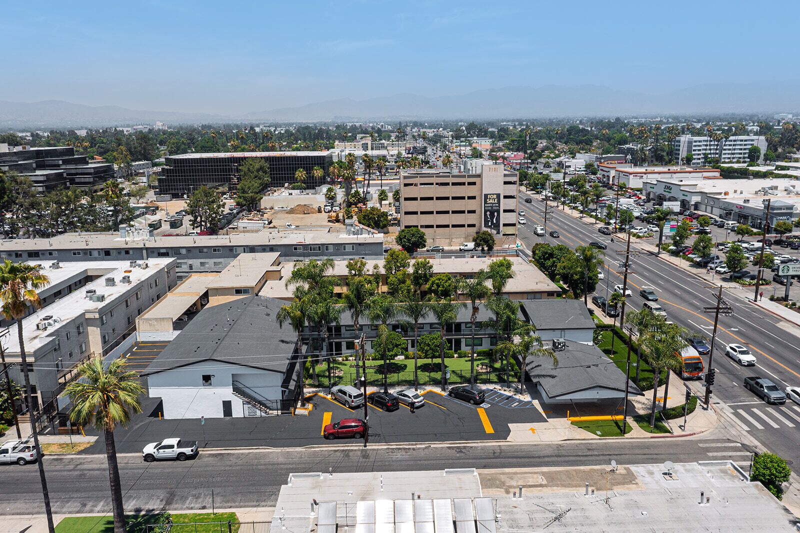 A multi-story parking garage with a 'For Sale' sign, surrounded by urban buildings and palm trees.