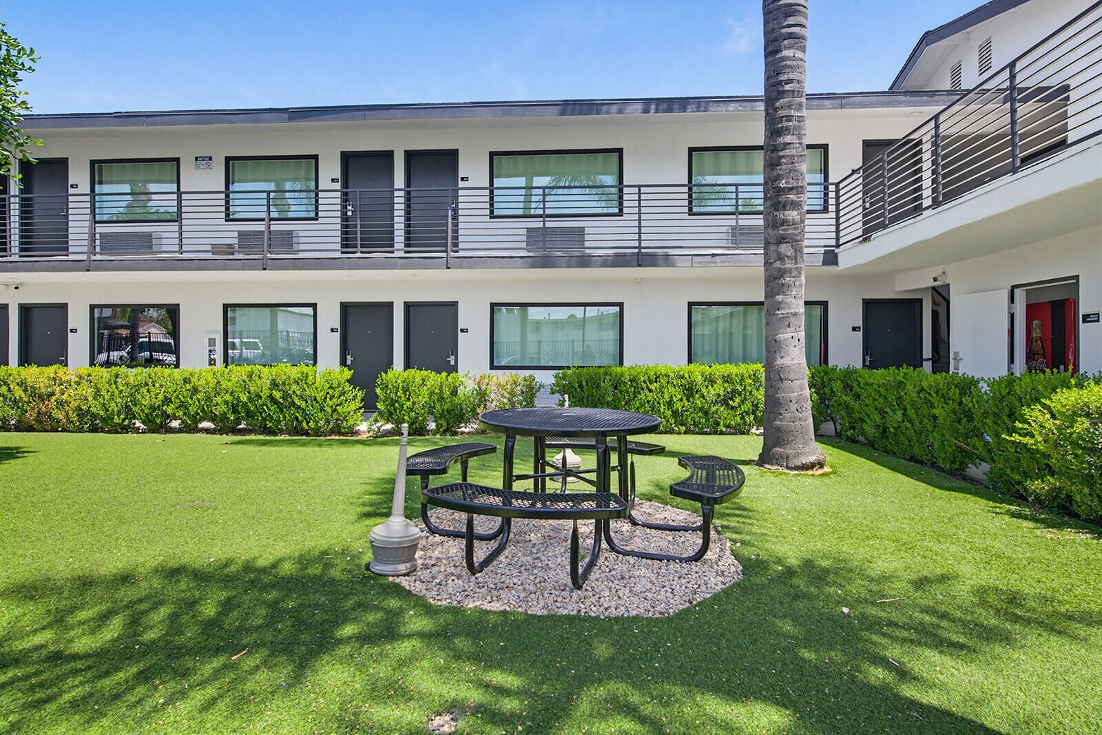 A modern building with a courtyard featuring a round table and chairs, surrounded by well-maintained hedges and a palm tree.