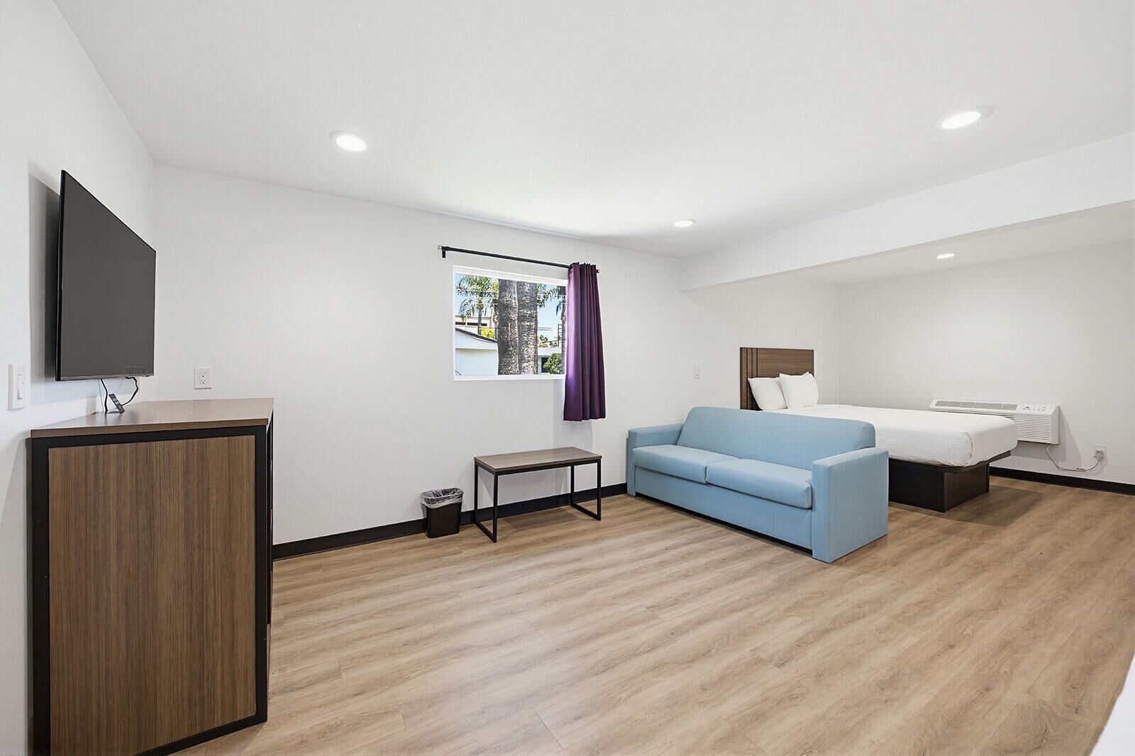 Ein Hotelzimmer mit einem Bett, einem blauen Sofa, einem kleinen Tisch, einem an einer Kommode befestigten Fernseher und einem Fenster mit Vorhängen.