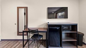 A compact room with a desk, a chair, a microwave, a trash bin, and a mirror on the wall.