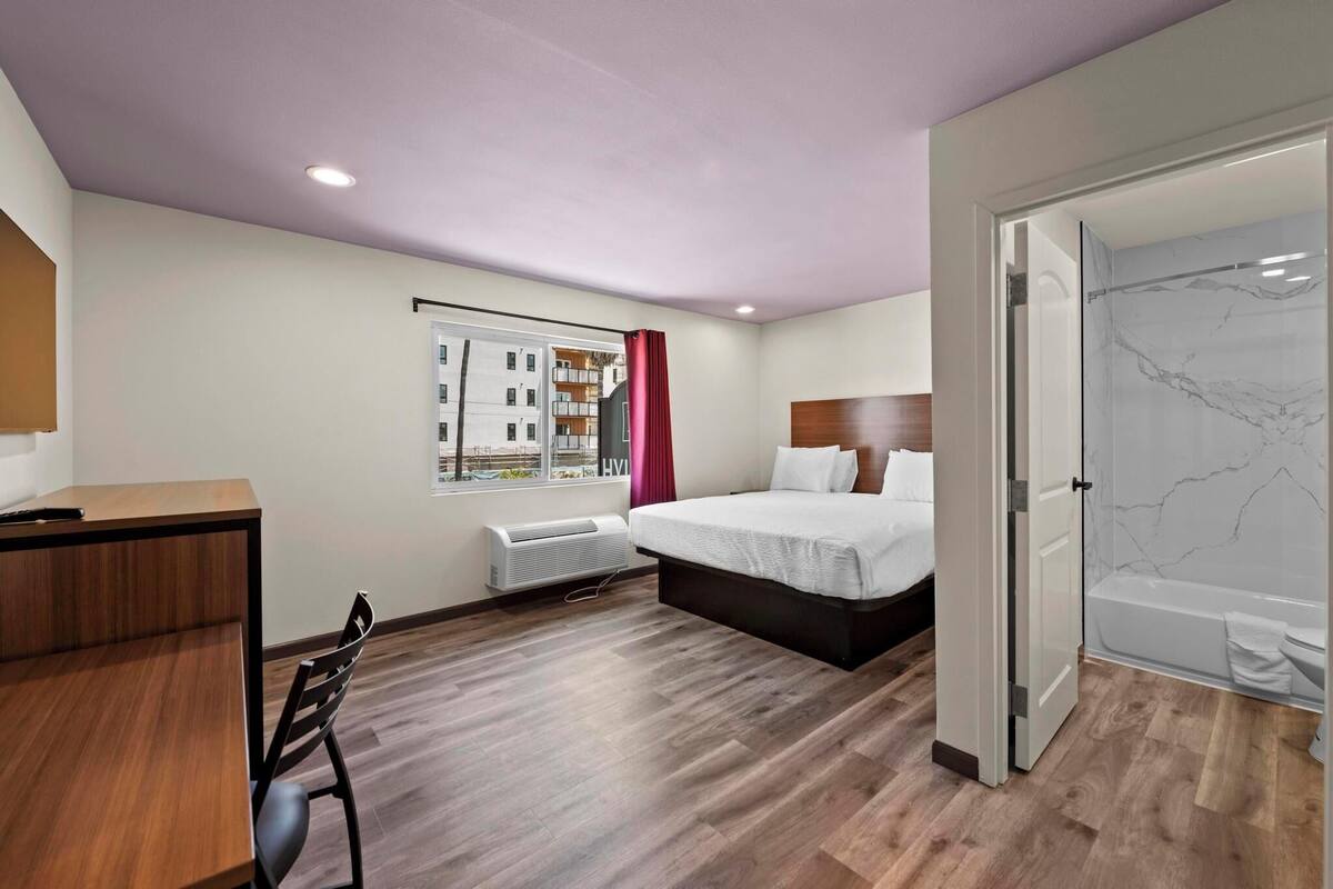 A hotel room with a bed, a desk, a chair, a window with a view of buildings, and a bathroom with a marble wall.