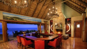 A spacious dining area with a large wooden table and red chairs, a stone fireplace, and a view of the ocean.
