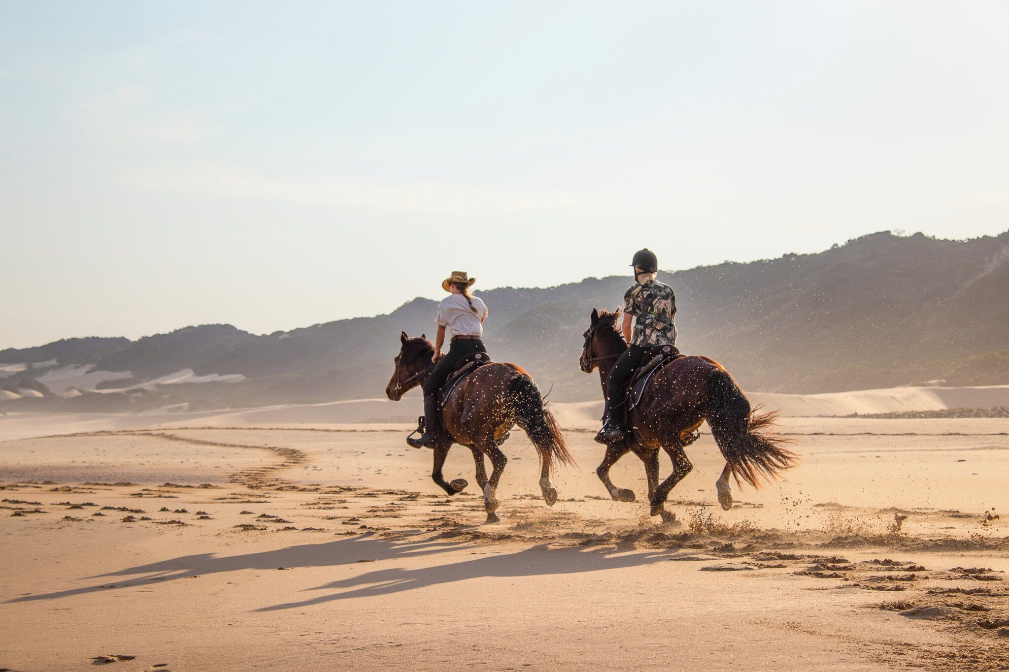 Dos personas montando a caballo en una playa arenosa, con colinas al fondo.