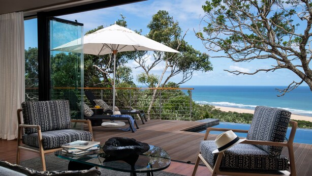 A modern living room with a glass wall overlooking the ocean, furnished with two patterned chairs, a glass coffee table, and a white umbrella.